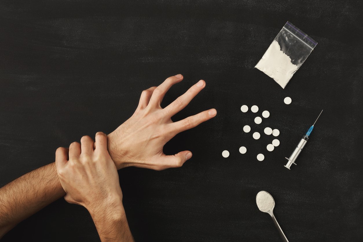 Man's hands trying to abstain from drugs on dark table with cooked heroine, pills, spoon and plastic bag. Concept for drug addiction and International Day against Drug Abuse, top view
